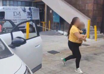 Fotografía de una mujer sosteniendo a un bebé entre brazos caminando hacia la puerta del hospital mientras a su lado hay un automovil estacionado con las puertas abiertas y una ambulancia a la esquina del edificio que se alcanza a notar como fondo