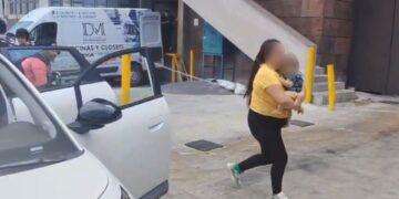 Fotografía de una mujer sosteniendo a un bebé entre brazos caminando hacia la puerta del hospital mientras a su lado hay un automovil estacionado con las puertas abiertas y una ambulancia a la esquina del edificio que se alcanza a notar como fondo