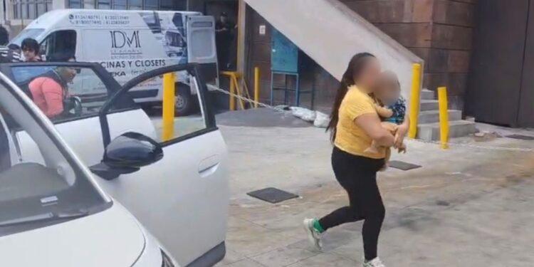 Fotografía de una mujer sosteniendo a un bebé entre brazos caminando hacia la puerta del hospital mientras a su lado hay un automovil estacionado con las puertas abiertas y una ambulancia a la esquina del edificio que se alcanza a notar como fondo